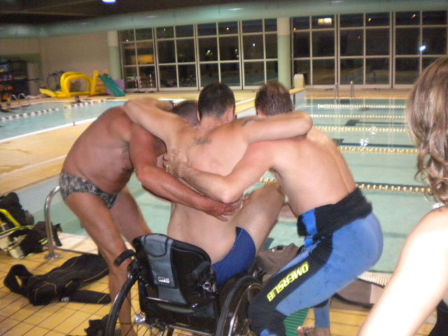 Uomo in sedia a rotelle viene aiutato ad entrare in piscina da due persone