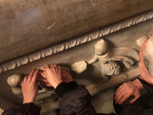 Mani in primo piano di una guida che accompagna un visitatore non vedente nella percezione tattile della simbologia cristiana presente su un sarcofago di epoca romana